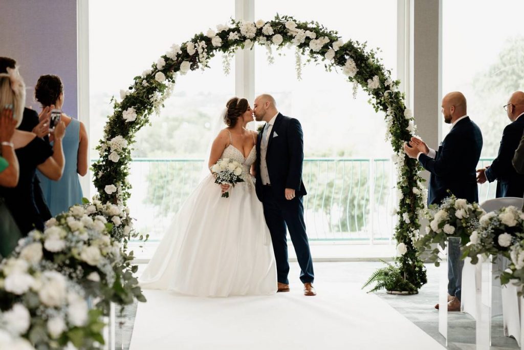 A just married couple kissing in front of their wedding party under a rose arch at the Crowne Plaza Marlow